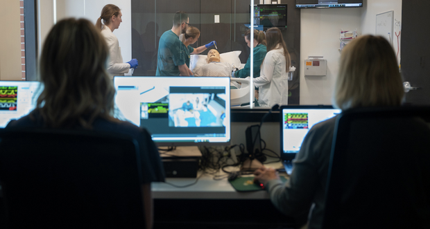 NDSU护理和药学专业的学生在模拟实验室中与正在经历蓝色代码的人体模型一起工作的照片, 他们的教授在控制室里观察并记录